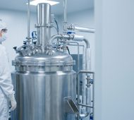 Scientist in protective white hazmat suit monitoring large stainless steel industrial chemical reactor and digital control panel in laboratory with copy space